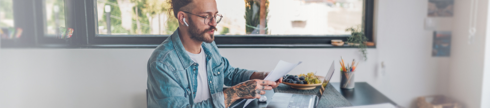 Man with headphones reading documents next to laptop.