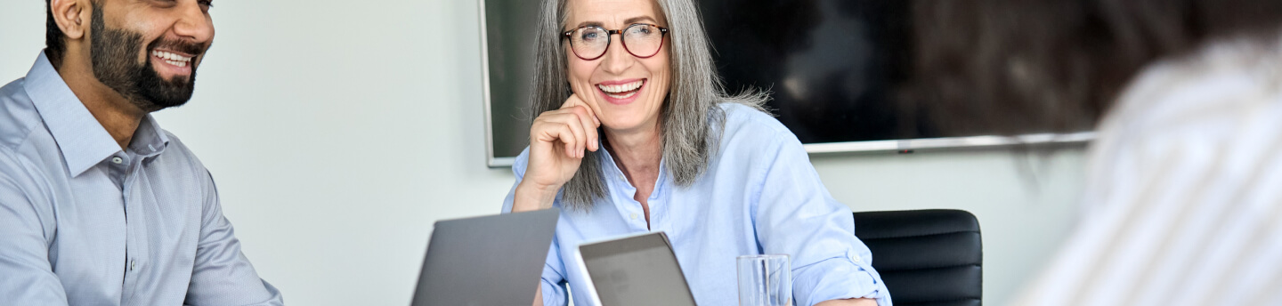 People in a business meeting, smiling older woman with glasses and laptop.
